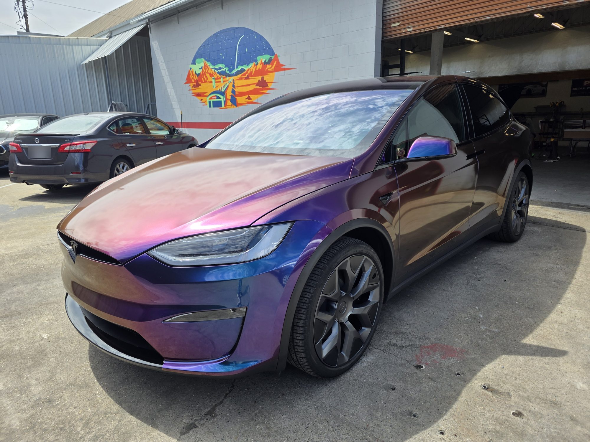 Tesla Model X in a purple-to-blue colour-flip wrap with full-body clear paint protection film over it, front three-quarter view by the mural at the Innovo shop in Oakland.