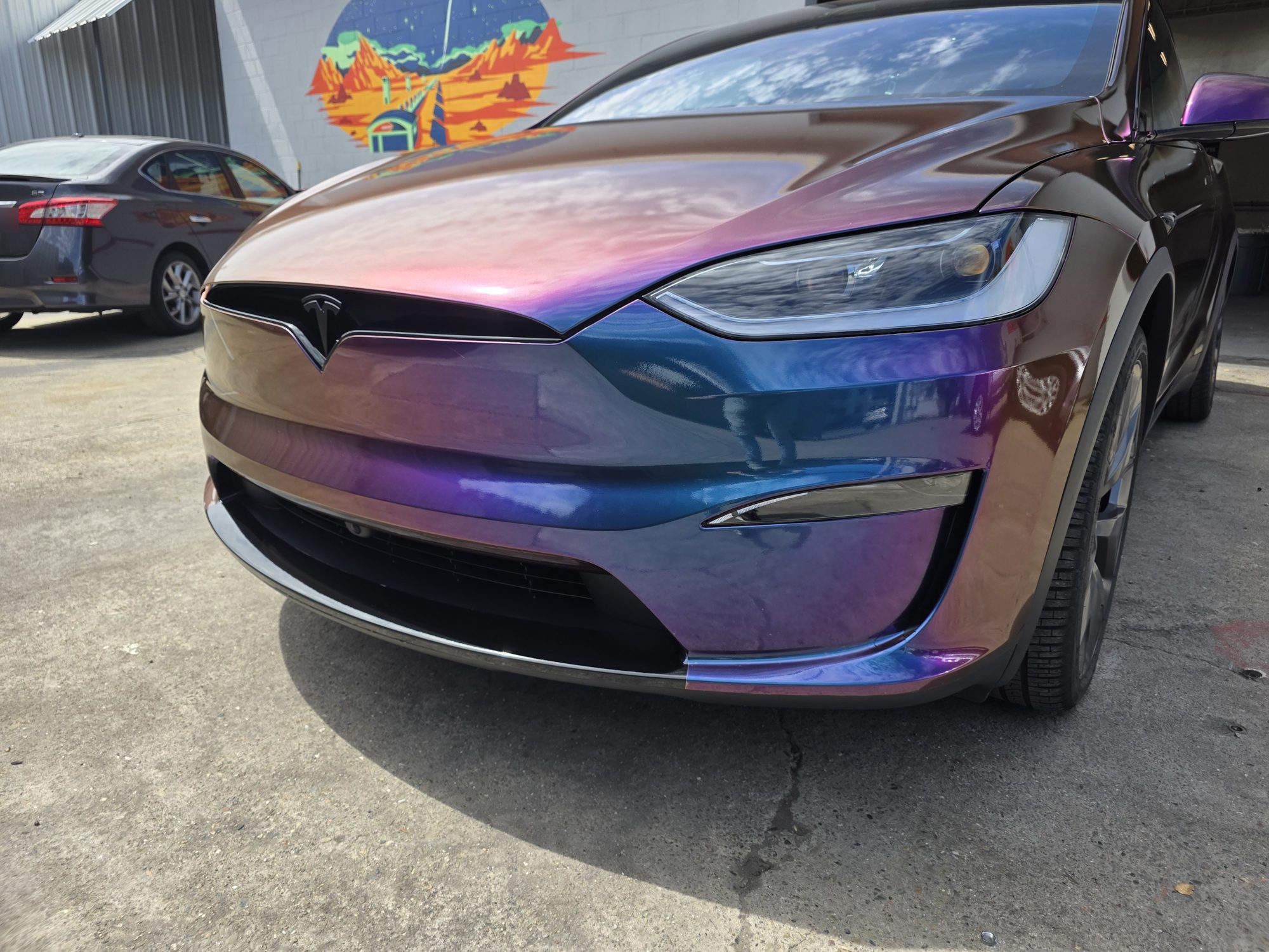 Nose and bumper detail on a Tesla Model X with clear paint protection film over a colour-flip wrap, Innovo shop mural behind.