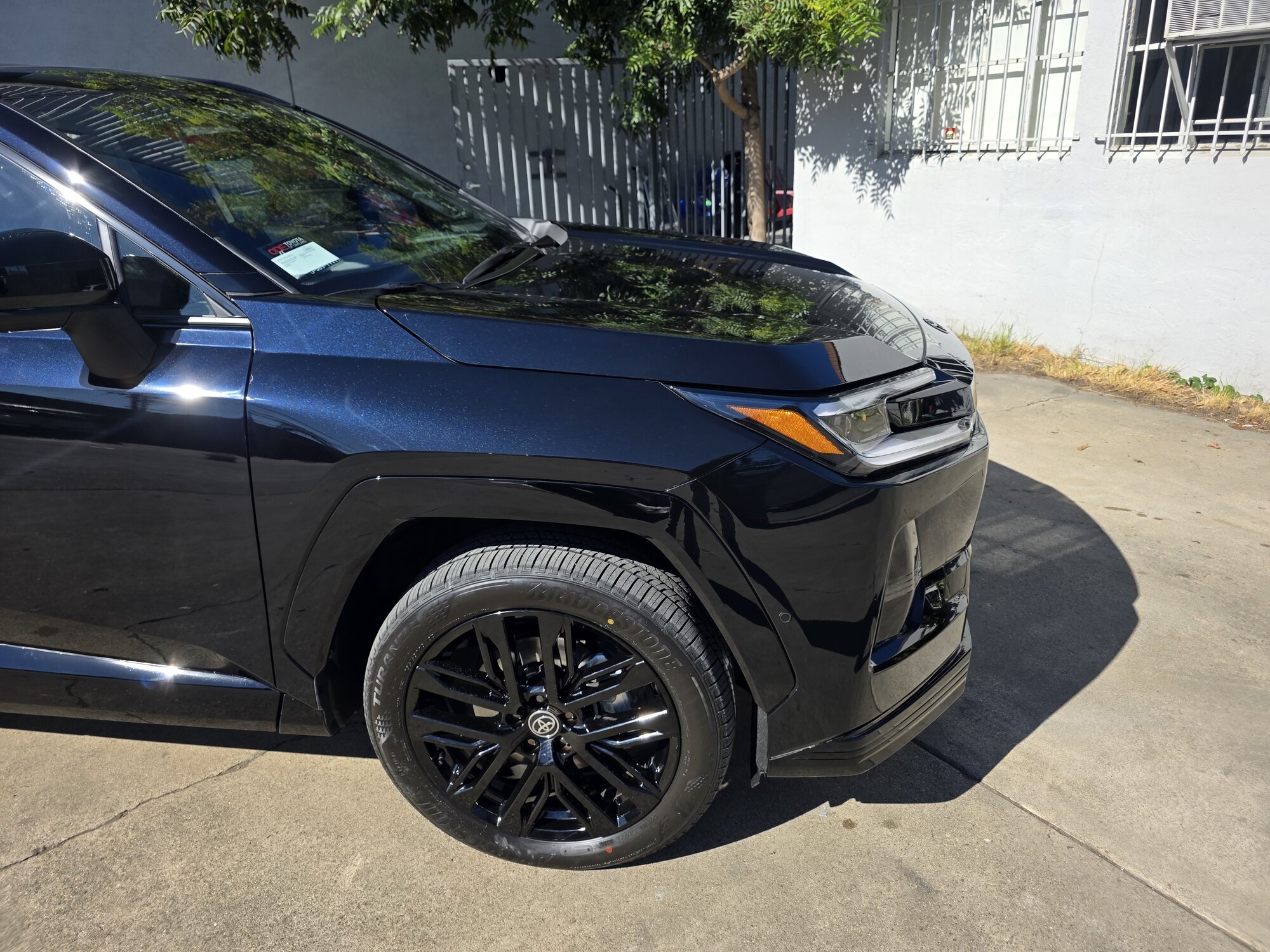 Front fender, headlight and wheel of a dark blue 2026 Toyota RAV4 after clear paint protection film, the metallic flake showing through the film in the sun.